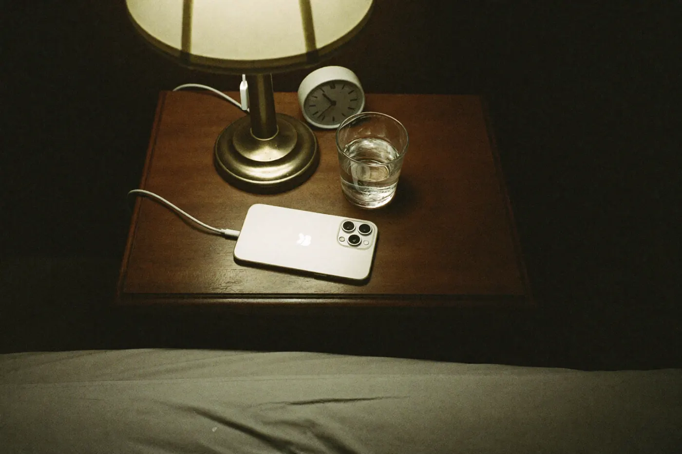A nightstand at night: a lamp, a glass of water, an alarm clock and a phone lying face down.